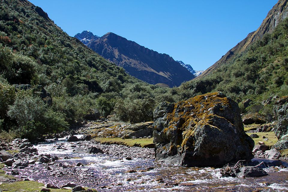 Mountainous landscape with a stream and large rock, under a clear blue sky.