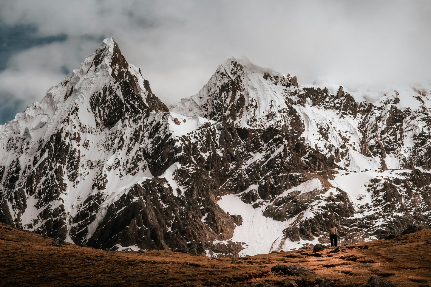 Snow-capped mountain range with a person standing at the base on a cloudy day.