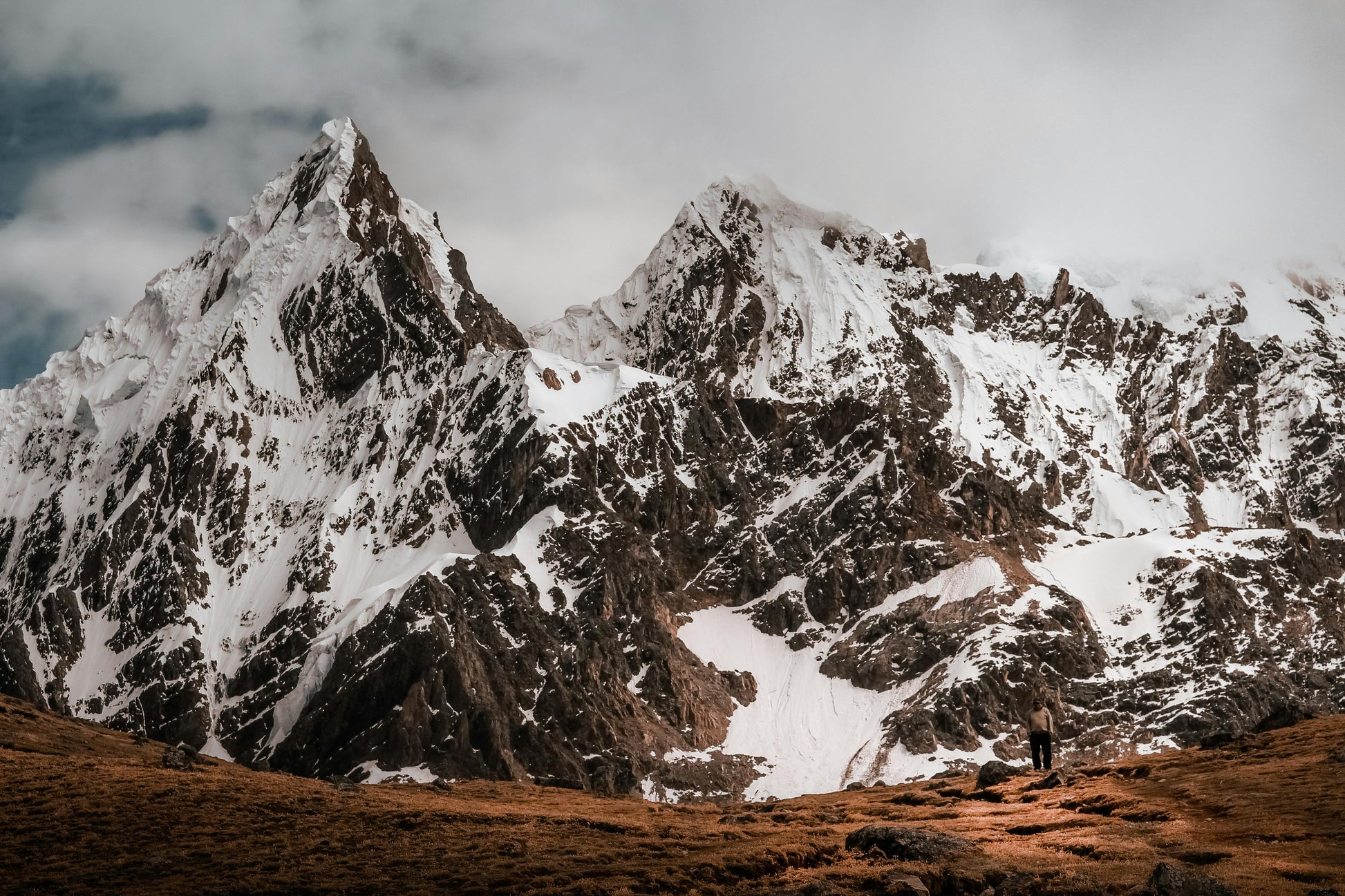 Snow-capped mountain range with a person standing at the base on a cloudy day.